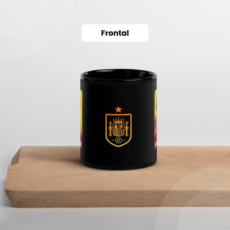 May include: Black ceramic mug with the Spanish coat of arms in gold and red. The mug has a star above the emblem and the word "Frontal" in white. The mug is on a wooden surface.