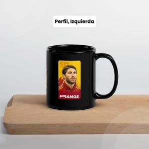 May include: A black ceramic mug with a portrait of a footballer in a red and yellow uniform. The mug has the text "RAMOS" and the number "15" on a yellow background. The text "Perfil, Izquierda" is above the mug.