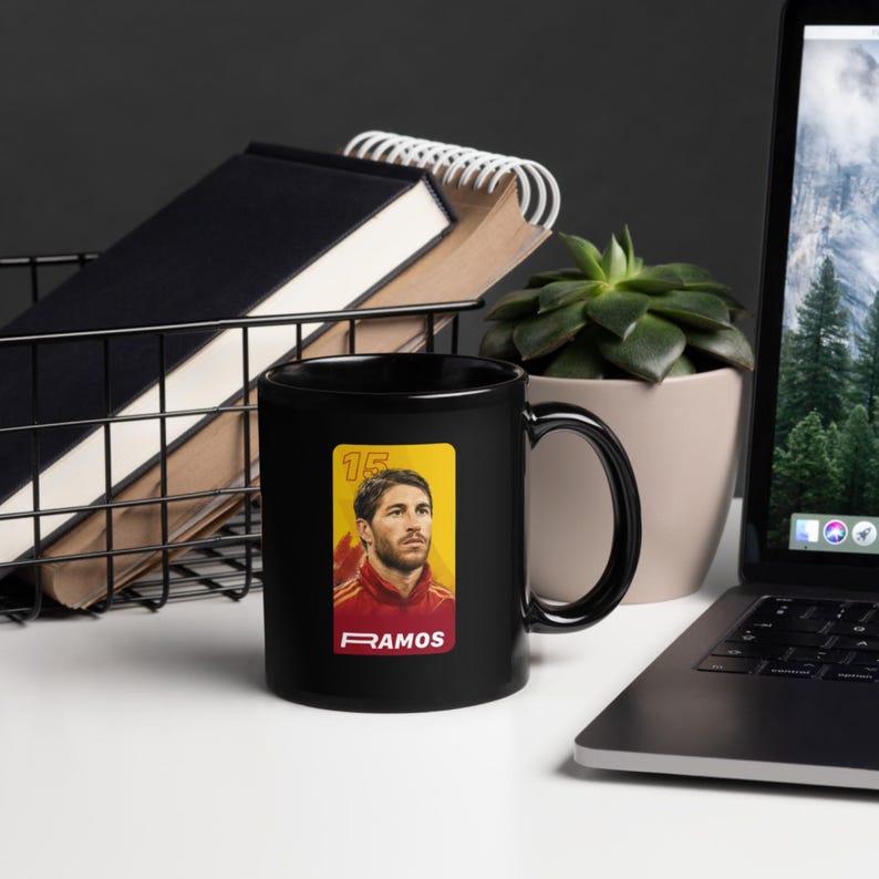 May include: A black coffee mug with a portrait of a soccer player, number 15, and the name RAMOS. The portrait is on a yellow background. The mug is on a white surface.