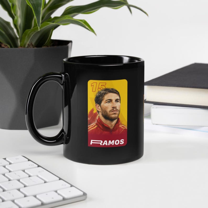May include: A black ceramic coffee mug with a portrait of a man in a red and yellow jersey. The mug has a black handle and the word "RAMOS" is printed on the bottom of the portrait.