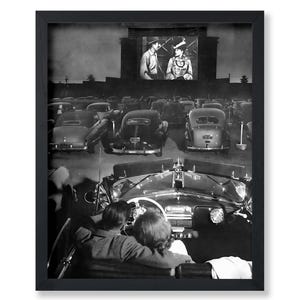 May include: Black and white framed print depicting a drive-in movie scene. A couple is seen in a classic car, watching a film on a large screen. Vintage cars fill the parking lot, creating a nostalgic atmosphere.