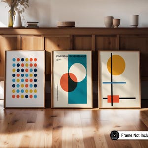 May include: Three framed art prints with geometric designs. One features a grid of colorful dots, another has overlapping circles and text, and the third showcases a yellow circle, black lines, and a red square. The frames are a light brown color.