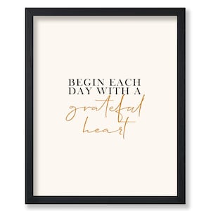 May include: A framed print with a black frame. The print has a cream-colored background with the words "BEGIN EACH DAY WITH A grateful heart" in black and gold script.