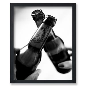 May include: Black and white framed print featuring two dark glass beer bottles clinking together. The bottles are filled with a dark liquid and have foam near the top. The image is a close-up shot, with the bottles in focus.