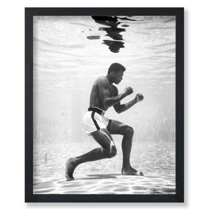 May include: Black and white framed print of a boxer underwater, in a fighting stance. The boxer is wearing white shorts with a black stripe and a black belt. The image is in a black frame.