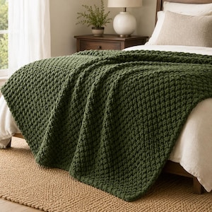 May include: A chunky knit throw blanket in a deep forest green color with a thick, textured weave pattern. The blanket is draped over a bed with white bedding, featuring a visible cable-like stitch construction throughout the entire fabric surface.