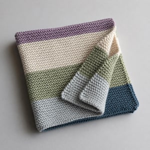 May include: A folded, knitted blanket with horizontal stripes in shades of purple, cream, green, light blue, and navy blue. The blanket is made of a soft, textured material and is folded over to reveal the underside.