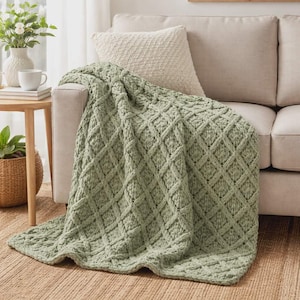 Può includere: Una coperta lavorata a maglia verde salvia con un motivo a rombi è drappeggiata su un divano beige. La coperta è realizzata in un materiale morbido e strutturato. Un cuscino beige poggia contro lo schienale del divano. È visibile un tavolino con una pianta e un vaso.