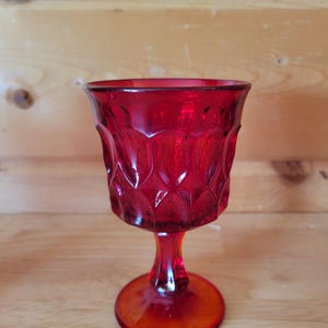 May include: A ruby red vintage goblet with a patterned design on the bowl. The goblet has a round base, a stem, and a wide rim. The glass is transparent, allowing light to pass through. The goblet is likely made of glass.