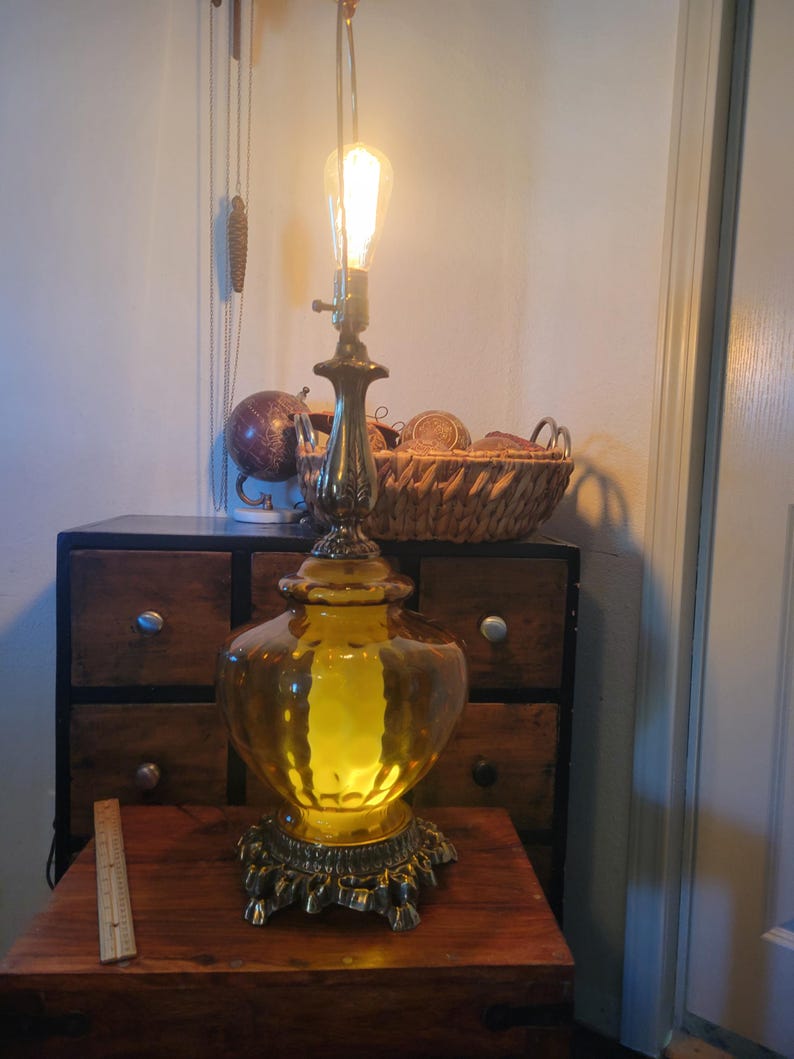 Może przedstawiać: Antyczna lampa stołowa z ż&oacute;łtą szklaną podstawą i mosiężnymi akcentami. Lampa posiada żar&oacute;wkę w stylu Edisona i stoi na drewnianym stole. Na stole widoczna jest linijka. To element wystroju wnętrz w stylu vintage.