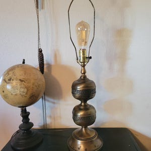 Może przedstawiać: Wysoka lampa stołowa z mosiądzu w stylu antycznym z dwupoziomową, zaokrągloną podstawą z wytłoczonymi wzorami kwiatowymi. Lampa posiada metalowy stelaż oraz pojedynczą żarówkę typu Edison z widocznym żarnikiem w górnej części.