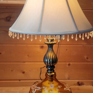 May include: An amber-colored table lamp with a cream-colored lampshade. The lamp features a decorative floral design on the base and a beaded trim. The lamp is made of metal and glass.
