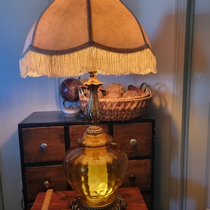 Może przedstawiać: Antyczna lampa stołowa z dużą, ozdobną podstawą ze szkła w kolorze bursztynu i abażurem z frędzlami. Lampa ma podstawę i akcenty z metalu w kolorze mosiądzu. Klosz jest w jasnobrązowym kolorze z ciemniejszym wykończeniem.