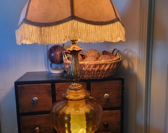 May include: An antique table lamp with a large, ornate amber glass base and a fringed lampshade. The lamp has a brass-colored metal base and accents. The lampshade is a light tan color with a darker trim.