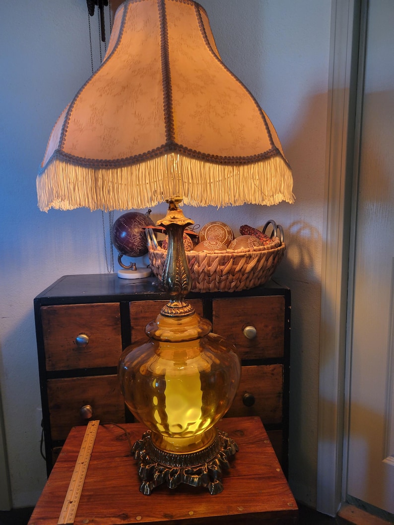 Może przedstawiać: Antyczna lampa stołowa z dużą, ozdobną podstawą ze szkła w kolorze bursztynu i abażurem z frędzlami. Lampa ma podstawę i akcenty z metalu w kolorze mosiądzu. Klosz jest w jasnobrązowym kolorze z ciemniejszym wykończeniem.
