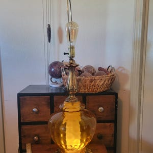 Może przedstawiać: Zabytkowa szklana lampa stołowa w kolorze bursztynu z mosiężną podstawą i akcentami. Lampa posiada duży, zaokrąglony szklany klosz i ozdobny mosiężny trzon. Żar&oacute;wka jest widoczna, a lampa jest włączona. Lampa ma około 71 cm wysokości.