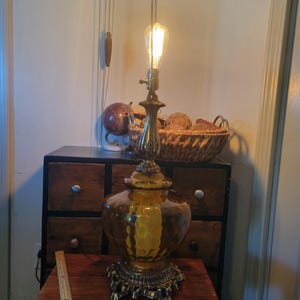 Może przedstawiać: Antyczna lampa stołowa z ozdobną mosiężną podstawą i dużym, bursztynowym szklanym kloszem. Lampa posiada żar&oacute;wkę w stylu Edisona i stoi na drewnianym stole. Obok lampy znajduje się linijka. Zabytkowy element wystroju wnętrz.