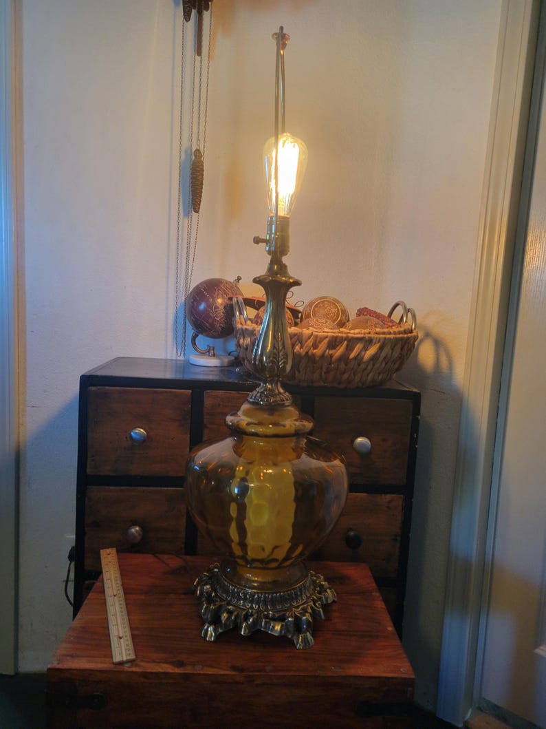 Może przedstawiać: Antyczna lampa stołowa z ozdobną mosiężną podstawą i dużym, bursztynowym szklanym kloszem. Lampa posiada żar&oacute;wkę w stylu Edisona i stoi na drewnianym stole. Obok lampy znajduje się linijka. Zabytkowy element wystroju wnętrz.