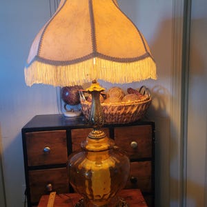 Może przedstawiać: Antyczna lampa stołowa z dużym, kremowym abażurem obszytym frędzlami. Lampa ma złotą, bursztynową szklaną podstawę i ozdobne mosiężne akcenty. Lampa stoi na drewnianym stole.