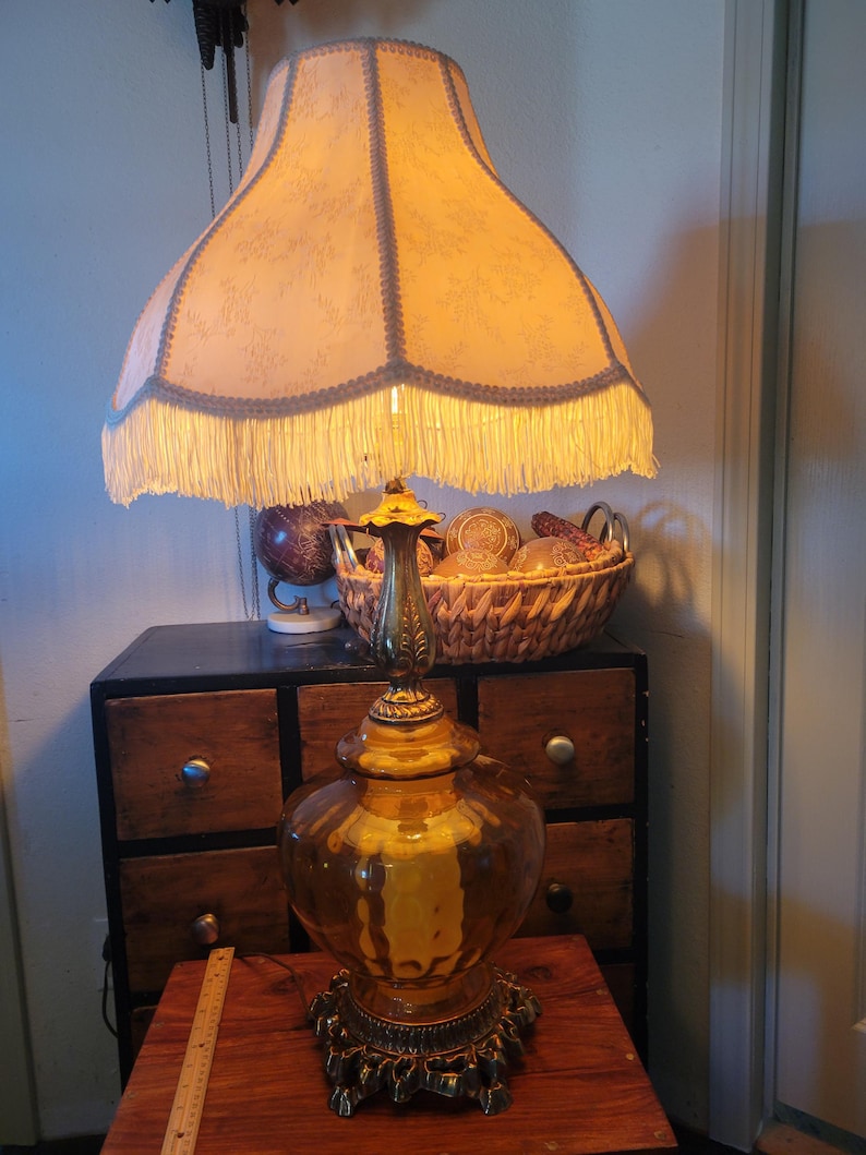 Może przedstawiać: Antyczna lampa stołowa z dużym, kremowym abażurem obszytym frędzlami. Lampa ma złotą, bursztynową szklaną podstawę i ozdobne mosiężne akcenty. Lampa stoi na drewnianym stole.
