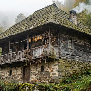 May include: A rustic, two-story wooden cabin with a stone foundation and a moss-covered roof. The cabin has a balcony, a chimney with smoke, and dried corn hanging from the balcony. The surrounding area is lush with green foliage.