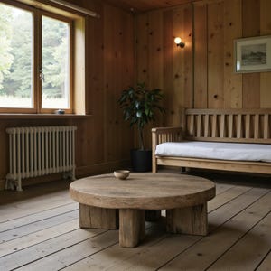 May include: A rustic, round wooden coffee table with a natural finish. The table has a thick, circular top supported by four sturdy, rectangular legs. The room features wooden paneling, a window, a bench, and a potted plant, creating a warm, inviting atmosphere.