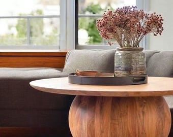 May include: A wooden coffee table with a round top and a large, rounded base. The table has a dark brown finish and is decorated with a black tray and a vase of dried flowers.