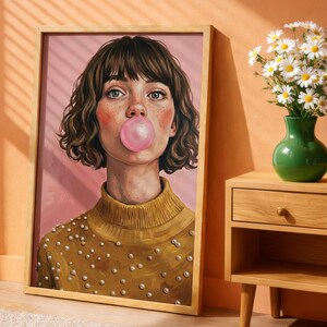 Peut inclure: Un portrait peint et encadré représentant une personne aux cheveux bruns courts et aux taches de rousseur, portant un pull jaune moutarde orné de perles blanches. La personne fait une grosse bulle de chewing-gum rose sur fond rose.