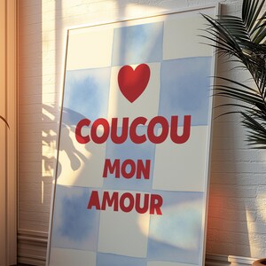 Peut inclure: Impression d'art encadrée avec un cœur rouge et le texte français "COUCOU MON AMOUR" en rouge. Le fond présente un motif à carreaux de carrés blancs et bleu clair. L'œuvre est affichée contre un mur de briques blanches.