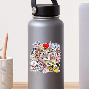 K&ouml;nnte beinhalten: Eine silberne Wasserflasche mit schwarzem Griff und Deckel. Die Flasche hat einen bunten Aufkleber mit Zeichentrickfiguren, Kassetten und floralen Elementen. Der Aufkleber enth&auml;lt Texte wie "Boy With Luv", "Butter" und "Fake Love".