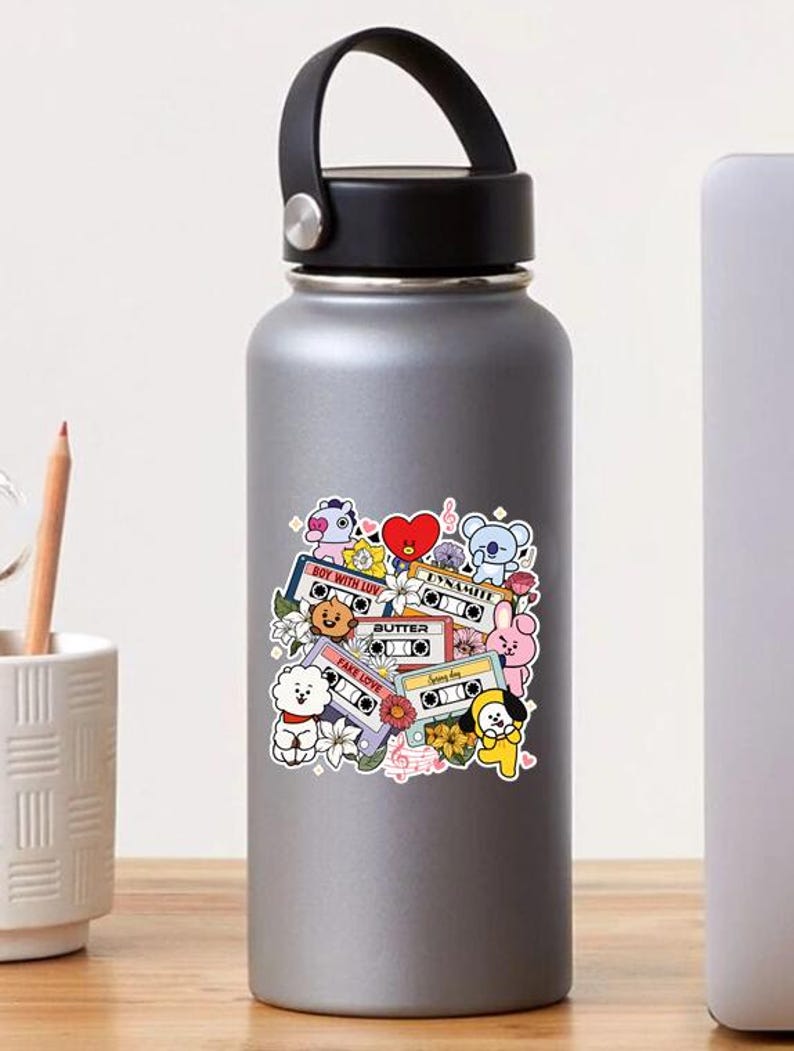 K&ouml;nnte beinhalten: Eine silberne Wasserflasche mit schwarzem Griff und Deckel. Die Flasche hat einen bunten Aufkleber mit Zeichentrickfiguren, Kassetten und floralen Elementen. Der Aufkleber enth&auml;lt Texte wie "Boy With Luv", "Butter" und "Fake Love".