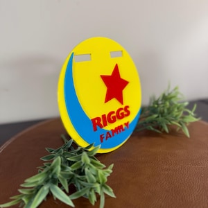 Peut inclure: Un panneau circulaire jaune, rouge et bleu avec le texte "RIGGS FAMILY" et une &eacute;toile rouge. Le panneau a une forme de croissant bleu et deux d&eacute;coupes rectangulaires en haut. Des brins verts sont plac&eacute;s autour du panneau.