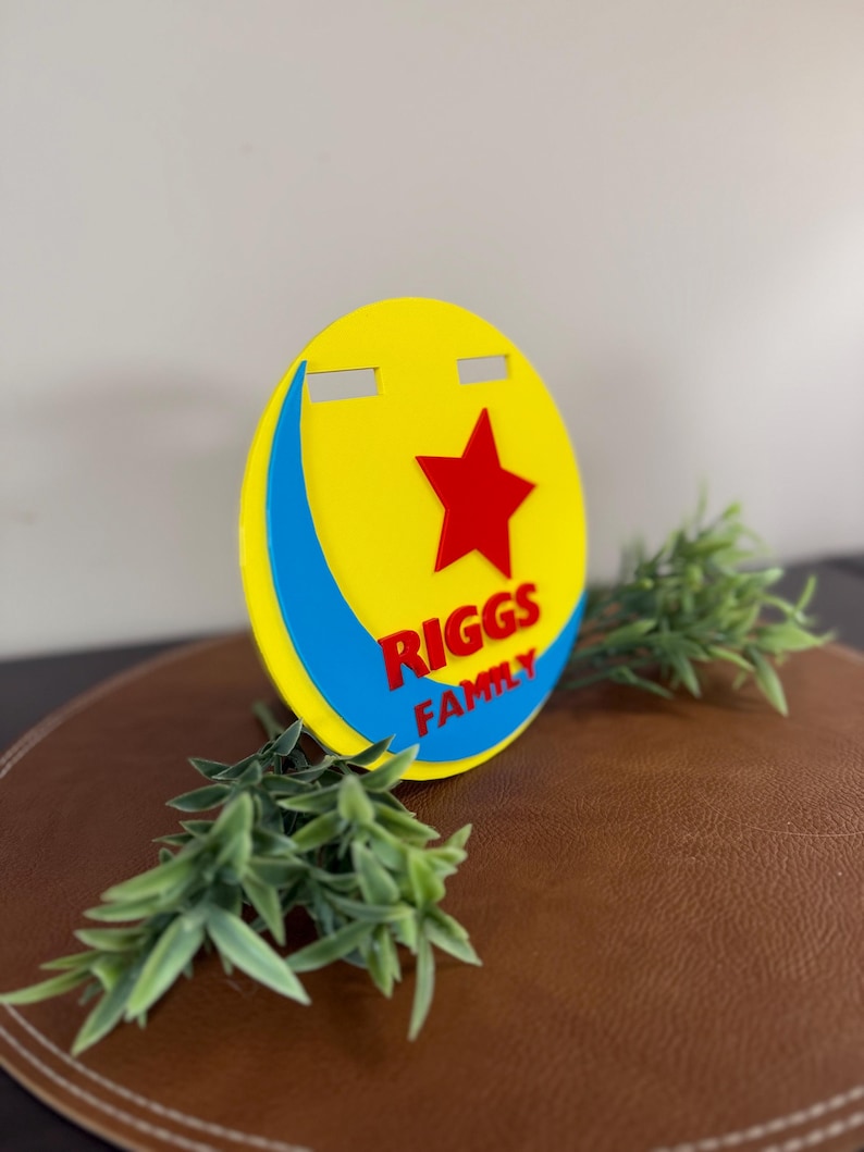 Peut inclure: Un panneau circulaire jaune, rouge et bleu avec le texte "RIGGS FAMILY" et une &eacute;toile rouge. Le panneau a une forme de croissant bleu et deux d&eacute;coupes rectangulaires en haut. Des brins verts sont plac&eacute;s autour du panneau.