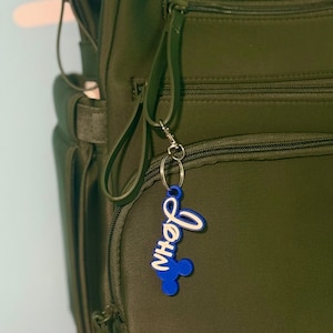 Puede incluir: Un llavero azul y blanco con el nombre "John" en fuente cursiva, sujeto a una mochila verde. El llavero tiene un diseño inspirado en Mickey Mouse. La mochila es verde oliva con múltiples compartimentos con cremallera.
