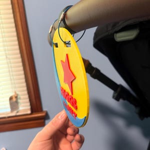 Peut inclure: Une &eacute;tiquette de poussette ovale jaune, bleue et rouge, avec le texte "RIGGS FAMILY" et une &eacute;toile rouge. L'&eacute;tiquette est attach&eacute;e &agrave; une poign&eacute;e de poussette avec un cordon bleu.