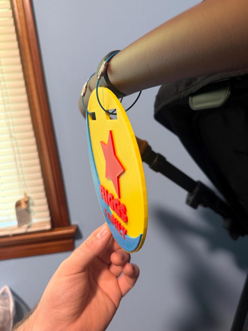 Peut inclure: Une &eacute;tiquette de poussette ovale jaune, bleue et rouge, avec le texte "RIGGS FAMILY" et une &eacute;toile rouge. L'&eacute;tiquette est attach&eacute;e &agrave; une poign&eacute;e de poussette avec un cordon bleu.