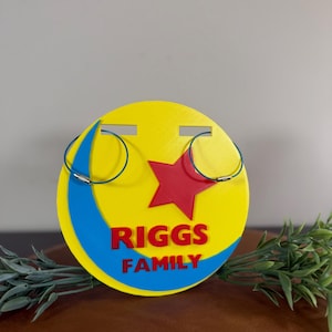 Peut inclure: Un pr&eacute;sentoir &agrave; boucles d'oreilles jaune et bleu avec une &eacute;toile rouge et le texte "RIGGS FAMILY". Deux paires de boucles d'oreilles cr&eacute;oles bleues sont affich&eacute;es sur le pr&eacute;sentoir. Le pr&eacute;sentoir est rond et pr&eacute;sente un motif de croissant de lune.