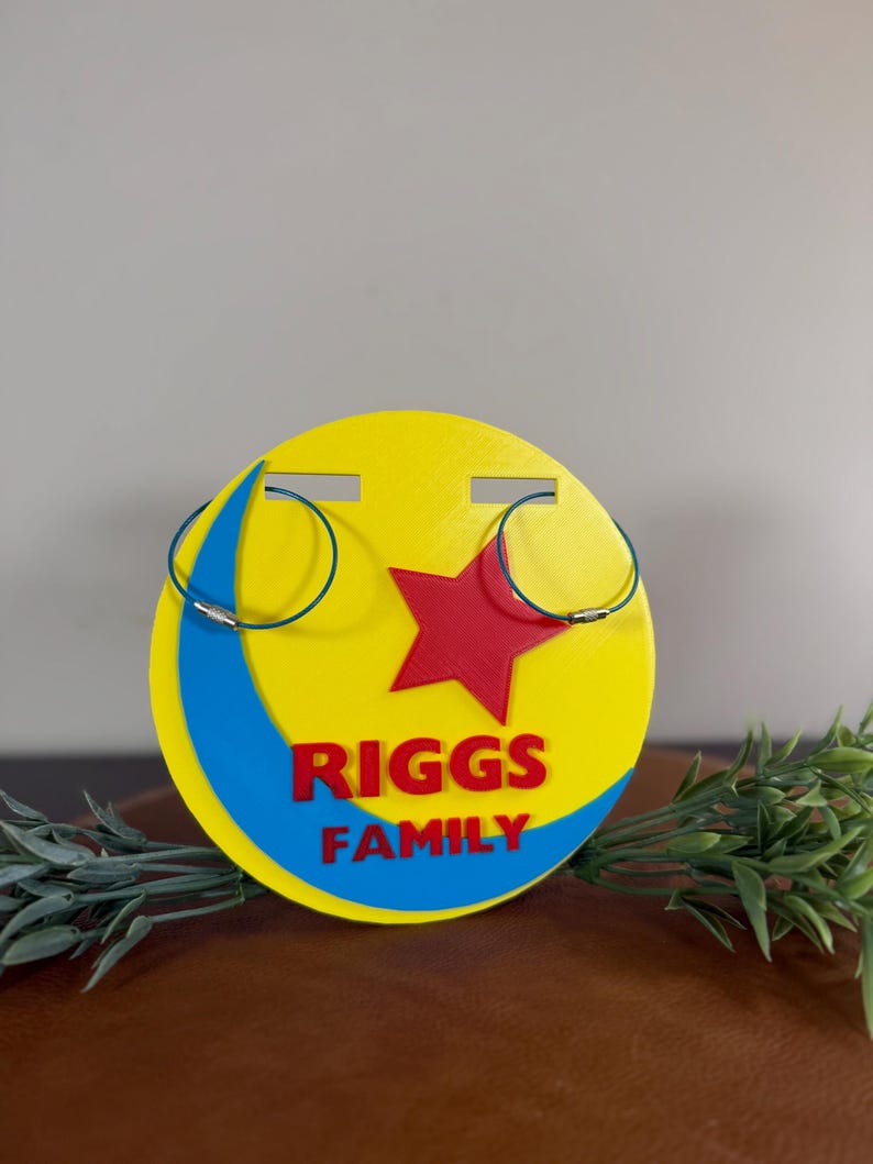 Peut inclure: Un pr&eacute;sentoir &agrave; boucles d'oreilles jaune et bleu avec une &eacute;toile rouge et le texte "RIGGS FAMILY". Deux paires de boucles d'oreilles cr&eacute;oles bleues sont affich&eacute;es sur le pr&eacute;sentoir. Le pr&eacute;sentoir est rond et pr&eacute;sente un motif de croissant de lune.