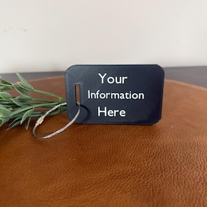 以下が含まれることがあります： 白字で「Your Information Here」と書かれた黒いラゲッジタグ。タグは角が丸い長方形で、取り付け用のスロットがあります。銀色のワイヤーループがタグに付いています。タグは茶色の表面にあります。