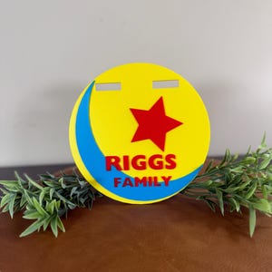 Peut inclure: Un panneau rond jaune avec une &eacute;toile rouge et un croissant bleu. Le texte "RIGGS FAMILY" est en rouge. Le panneau est pos&eacute; sur une surface marron avec du feuillage vert.