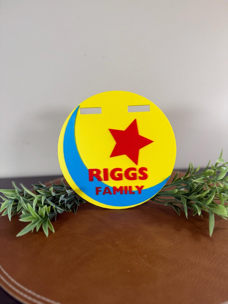 Peut inclure: Un panneau rond jaune avec une &eacute;toile rouge et un croissant bleu. Le texte "RIGGS FAMILY" est en rouge. Le panneau est pos&eacute; sur une surface marron avec du feuillage vert.