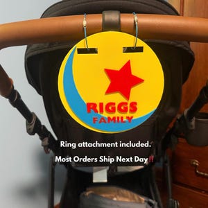 Peut inclure: Un porte-&eacute;tiquette de poussette circulaire jaune et bleu avec une &eacute;toile rouge et le texte "RIGGS FAMILY". L'&eacute;tiquette est attach&eacute;e &agrave; une poign&eacute;e de poussette. La poussette est noire avec des accents marron.