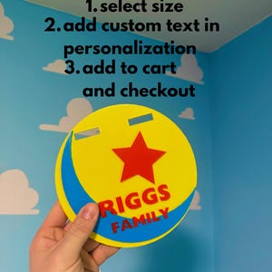 Peut inclure: Un panneau circulaire jaune et bleu avec une &eacute;toile rouge et le texte "RIGGS FAMILY". Le panneau comporte deux d&eacute;coupes rectangulaires et est tenu devant un mur bleu avec des d&eacute;calcomanies de nuages blancs. Le texte "HOW TO ORDER" et les instructions sont en haut.
