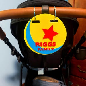 Peut inclure: Un panneau familial jaune, rouge et bleu avec le texte "RIGGS FAMILY" suspendu &agrave; une poign&eacute;e de poussette. Le panneau pr&eacute;sente une &eacute;toile rouge et un croissant de lune bleu.