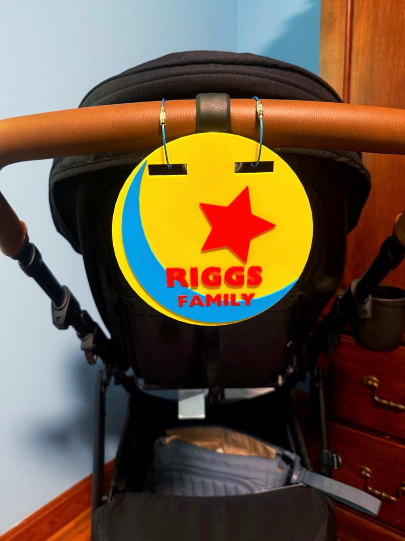 Peut inclure: Un panneau familial jaune, rouge et bleu avec le texte "RIGGS FAMILY" suspendu &agrave; une poign&eacute;e de poussette. Le panneau pr&eacute;sente une &eacute;toile rouge et un croissant de lune bleu.