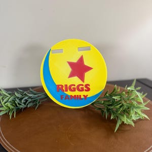 Peut inclure: Panneau circulaire jaune, rouge et bleu avec le texte "RIGGS FAMILY". Le panneau pr&eacute;sente une &eacute;toile rouge et un croissant bleu. Deux fentes horizontales sont d&eacute;coup&eacute;es en haut. Le panneau est sur une surface marron avec des accents de plantes vertes.