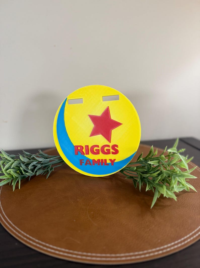 Peut inclure: Panneau circulaire jaune, rouge et bleu avec le texte "RIGGS FAMILY". Le panneau pr&eacute;sente une &eacute;toile rouge et un croissant bleu. Deux fentes horizontales sont d&eacute;coup&eacute;es en haut. Le panneau est sur une surface marron avec des accents de plantes vertes.