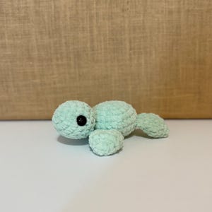 May include: A small, mint green crocheted turtle plush toy featuring a soft, textured yarn finish. The turtle has a rounded shell, a distinct head with a single visible black plastic safety eye, and small flippers extending from the sides.