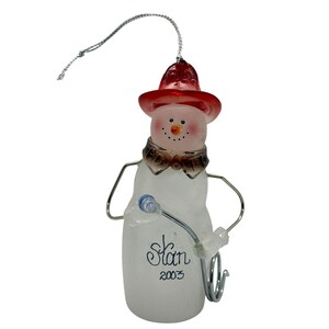 May include: A snowman Christmas ornament with a red hat, brown collar, and silver wire arms. The snowman has a carrot nose and a smiling face. The ornament has the name "Stan" and the year "2003" written on it. A festive holiday decoration.