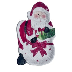 May include: White ceramic Santa Claus dish with a red hat, white beard, and a green and red gift. The dish has a pink and white bow and a white background with a textured pattern.